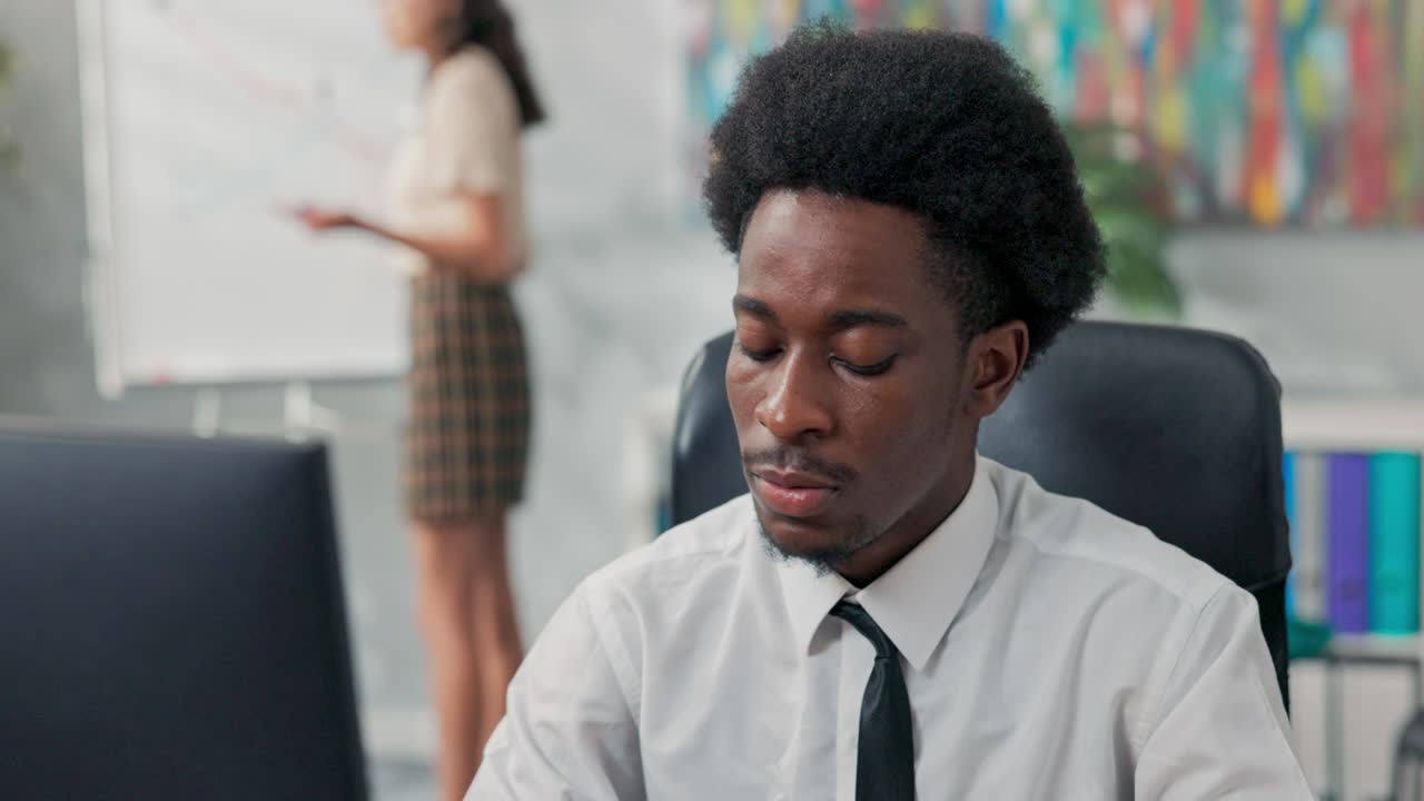 Un uomo in camicia bianca e cravatta con i capelli afro lavora concentrato al computer; sullo sfondo un collega sta lavorando a un progetto, creando statistiche disegnando un grafico.