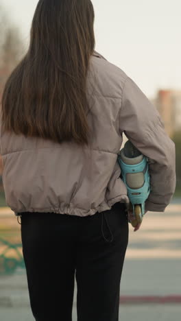 close-up de una chica con una chaqueta de melocotón y pantalones negros, sosteniendo una patineta turquesa, de pie en un camino del parque a principios de primavera con árboles desnudos y un fondo urbano borroso
