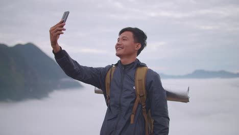 asian hiker male smiling and speaking to smartphone while live stream the view of top foggy mountain