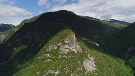 drone captures a steep, rocky mountain with lush greenery and a distant valley town on a sunny day.