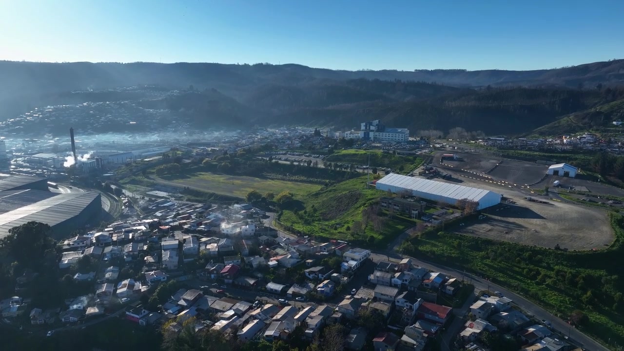 Voo avançando sobre uma cidade costeira nebulosa com edifícios industriais e bairros na encosta sob colinas ondulantes