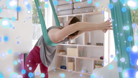 animation of blue and white light spots over caucasian woman practicing aerial yoga