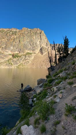 Vertical-View,-Young-Woman-Standing-on-Rock-by-Glacial-Alpine-Lake,-Sawtooth-Mountains,-Idaho-USA