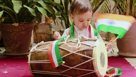 Niedliches-Kleinkind-Schwenkt-Indische-Trikolore-Flagge-Mit-Traditionellem-Dholak-Und-Tuch-Mit-Unschuldigem-Gesicht