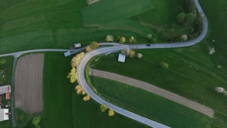 Drone's-eye-view:-Amidst-early-spring,-a-lone-car-ascends-a-winding-hillside-road,-unveiling-the-picturesque-landscape-around-the-journey,-with-the-lush-green-grass-with-a-golden-hue