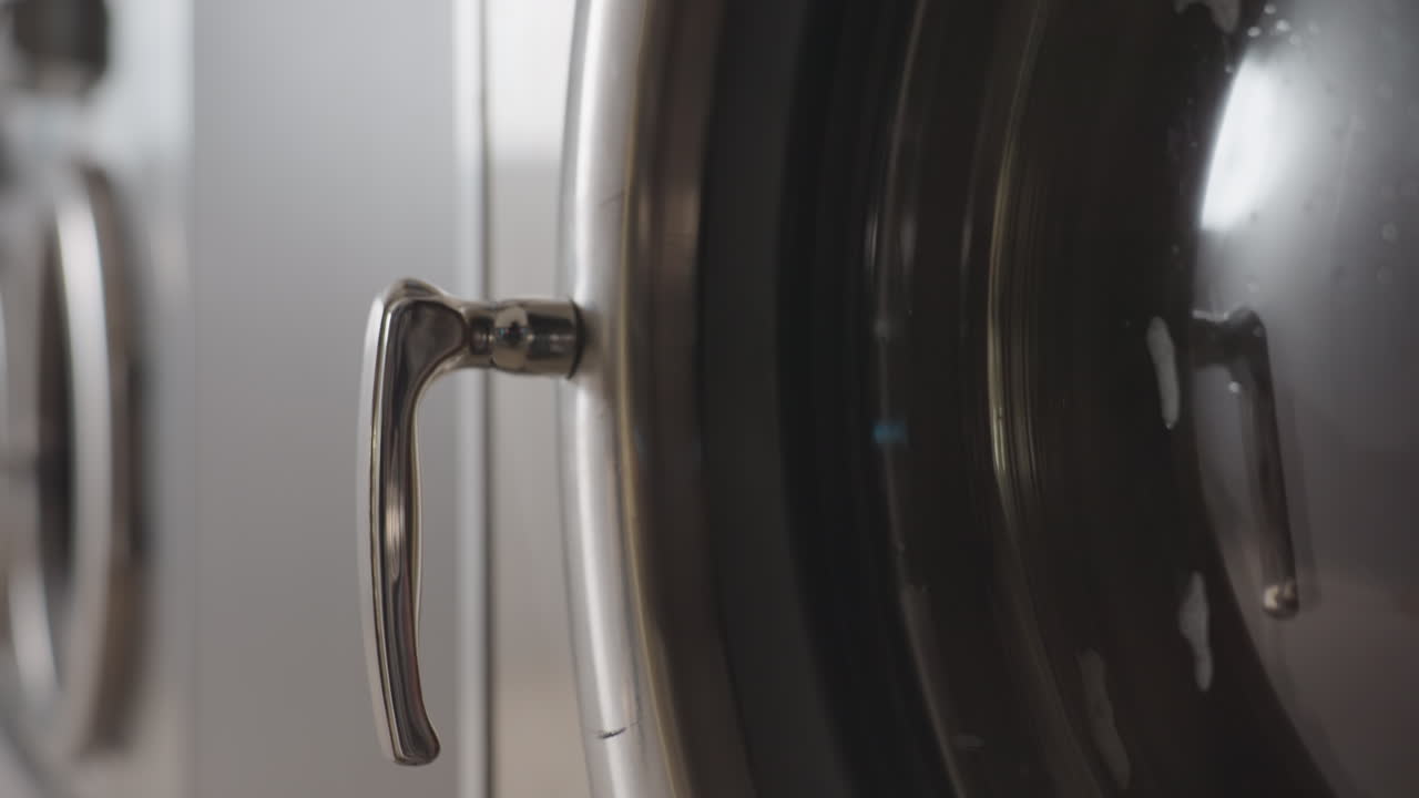 Downward view from number badge to door handle on stainless laundry machine while drum spins inside, soft reflection across metal surface, clean laundromat mood, motion with subtle bubbles on glass