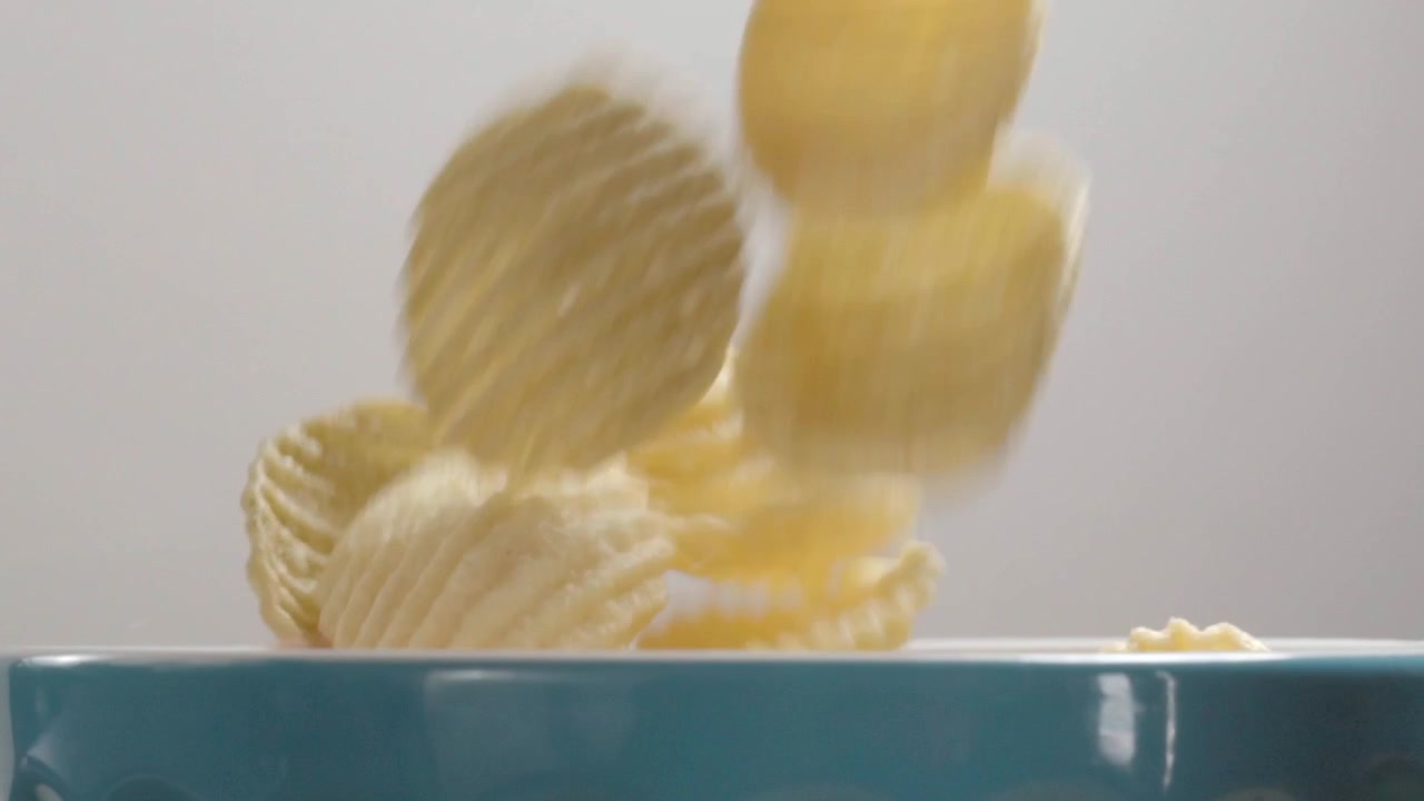 Slow motion close up of crispy ridged potato chips falling into a bowl, highlighting texture, crunch and snack food appeal against a clean background