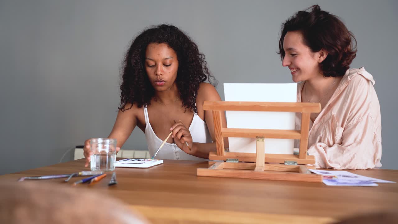 Focused diverse women sitting at table with painting brush and water colors