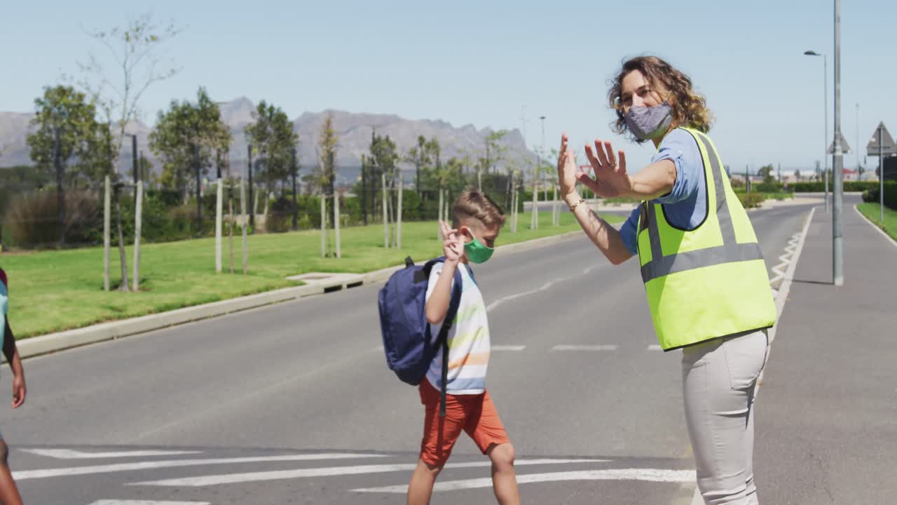 카우카시안 여성 교사가 얼굴 마스크와 높은 가시성 베스트를 착용하여 보행자에 대한 교통을 멈추고 있습니다.