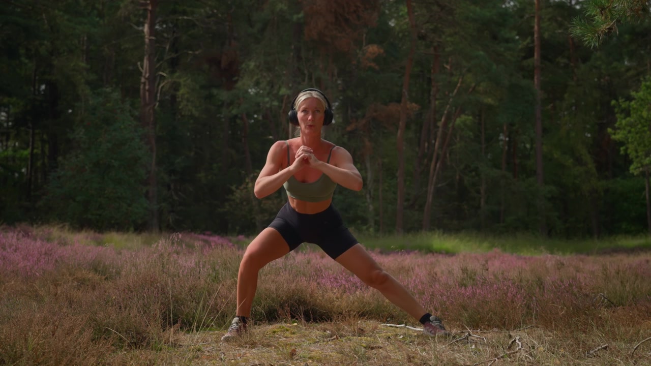 Riprese al rallentatore di una donna caucasica atletica in tenuta sportiva che esegue squat cosacchi nella natura con le cuffie, combinando esercizio all'aperto e allenamento della forza tra erica viola e bosco