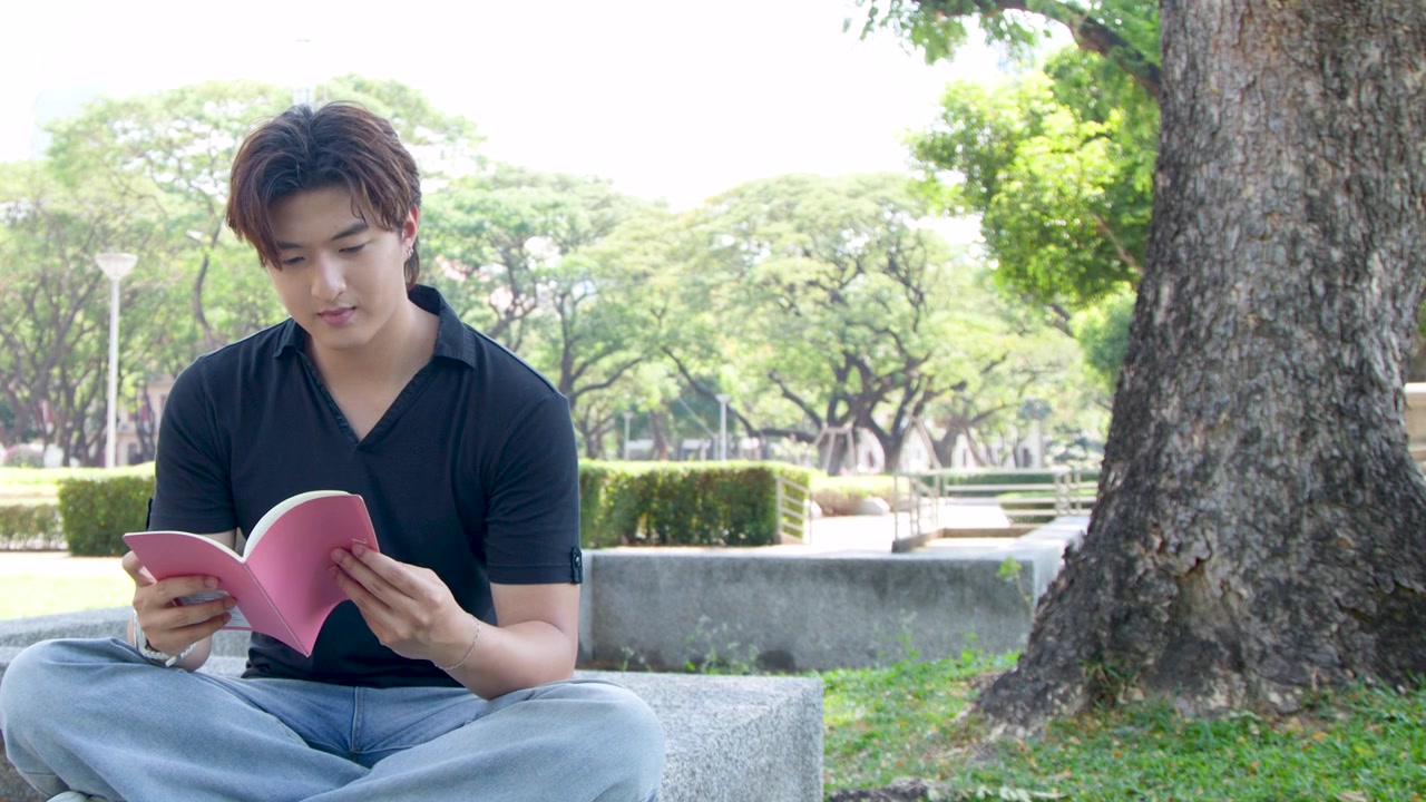 ชายหนุ่มชาวไทยนั่งขัดสมาธิกลางแจ้ง กำลังอ่านหนังสือสีชมพูใต้แสงแดดจ้า