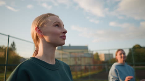 close-up of lady in green sweater playing volleyball with another lady in blue sweater blurred in the background, with a building and sunlight creating a warm glow