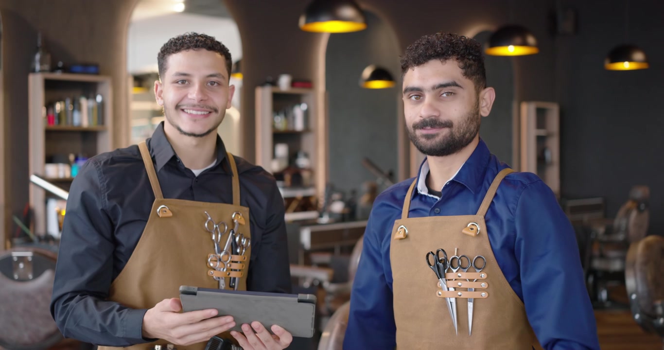 Dois barbeiros usando aventais com ferramentas, apontando, tocando no tablet e revisando conteúdo no salão
