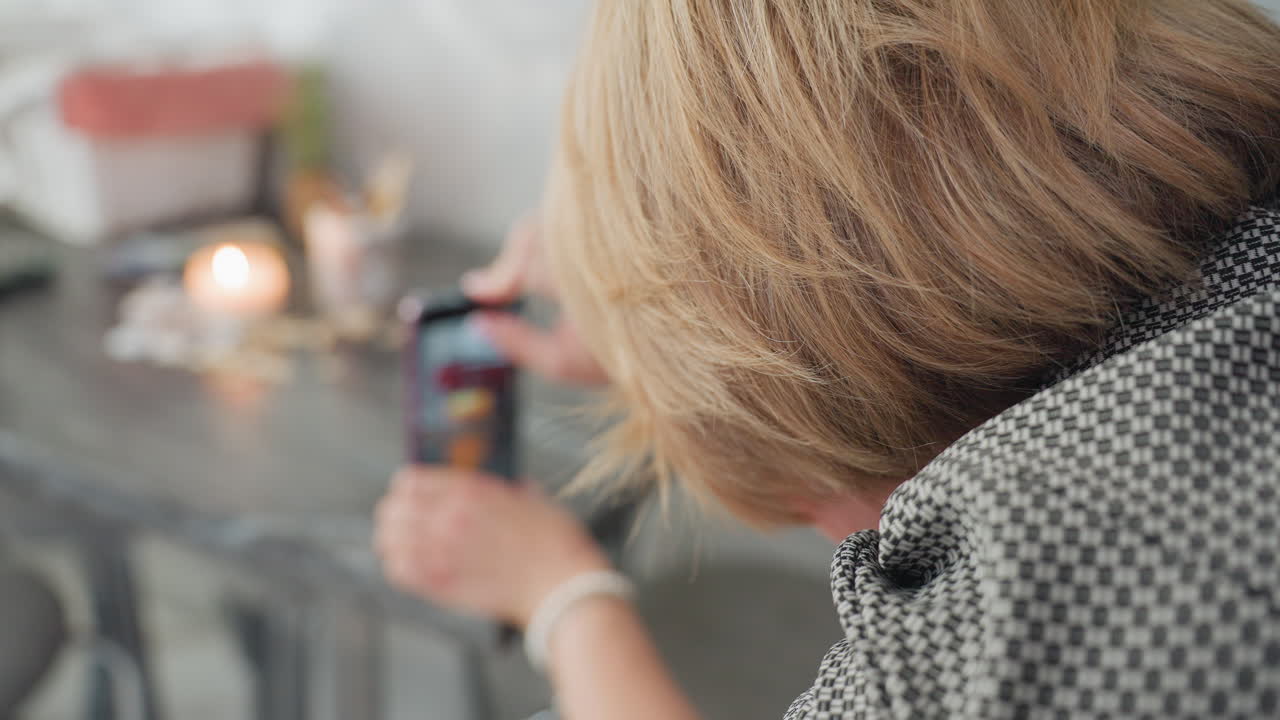 Close up rear view of young woman capturing candlelight setup with smartphone held upside down as she adjusts backward, creating warm peaceful atmosphere with seashells and candle