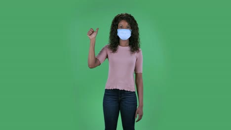 portrait of beautiful african american young woman in medical protective face mask is looking at camera and shows fist, then counting one, two, three, four, five fingers. burning brunette with wavy hair in jeans and a beige blouse on a green screen in the