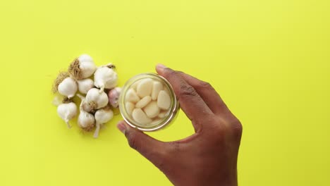 close-up view of a hand holding a jar of garlic cloves, with more garlic bulbs on a yellow background