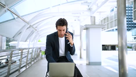 confident businessman using laptop working outdoors in the financial district in the city. using phone talking to client solving problems