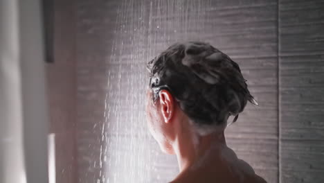 mujer con cabello espumoso bajo el agua de la ducha de primer plano. dama lava el champú de la cabeza relajándose en el baño. mujer cuida del cuerpo en el baño moderno