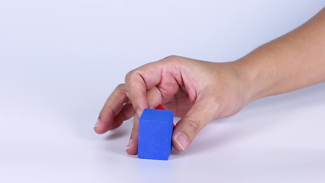 A person's hand carefully balances a red pyramid on top of a blue cube