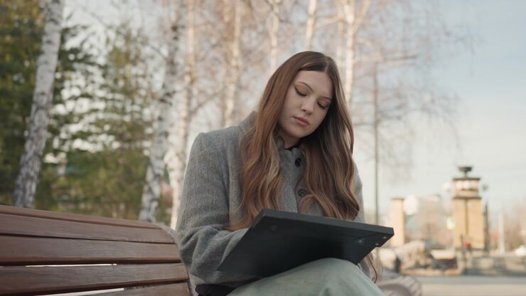 Kvinde tegner på tablet udendørs. Afslappet kunstner skitserer på tablet midt i naturlige omgivelser. Engageret kvinde illustrerer med stylus på tablet i fredfyldte udendørs omgivelser om vinteren.
