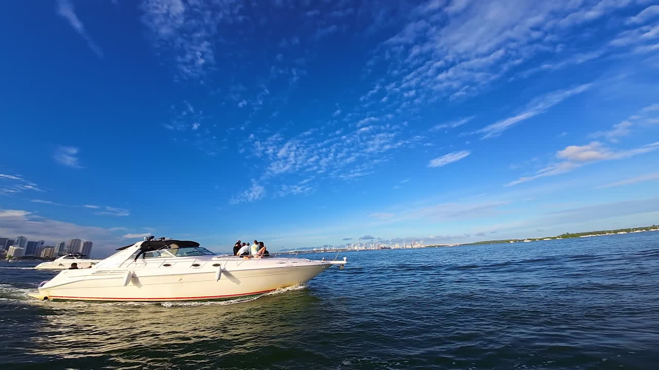 Approaching a motor boat with a group of young people on board. City skyline at backdrop. Low angle view at the blue sky with light cloudscape.