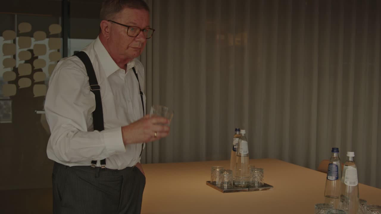 An older CEO pours himself a glass of water in his office and walks away