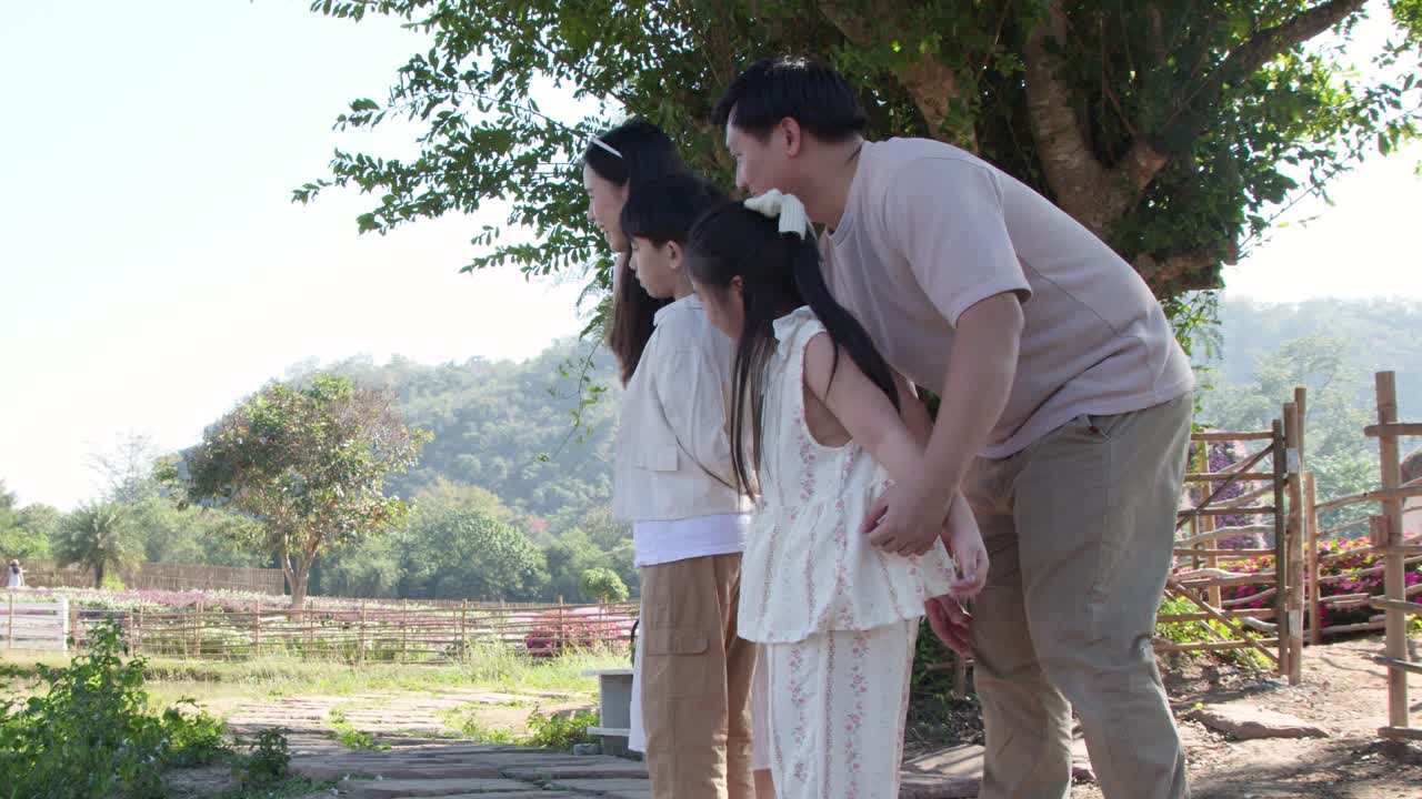 A happy Thai family stands together outdoors in a sunny garden with a bamboo fence