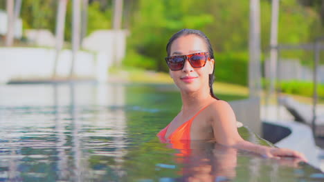 face close-up of young asian woman in her 30th inside the water leaning on the border of the infinity pool and turning sight into the camera with smile