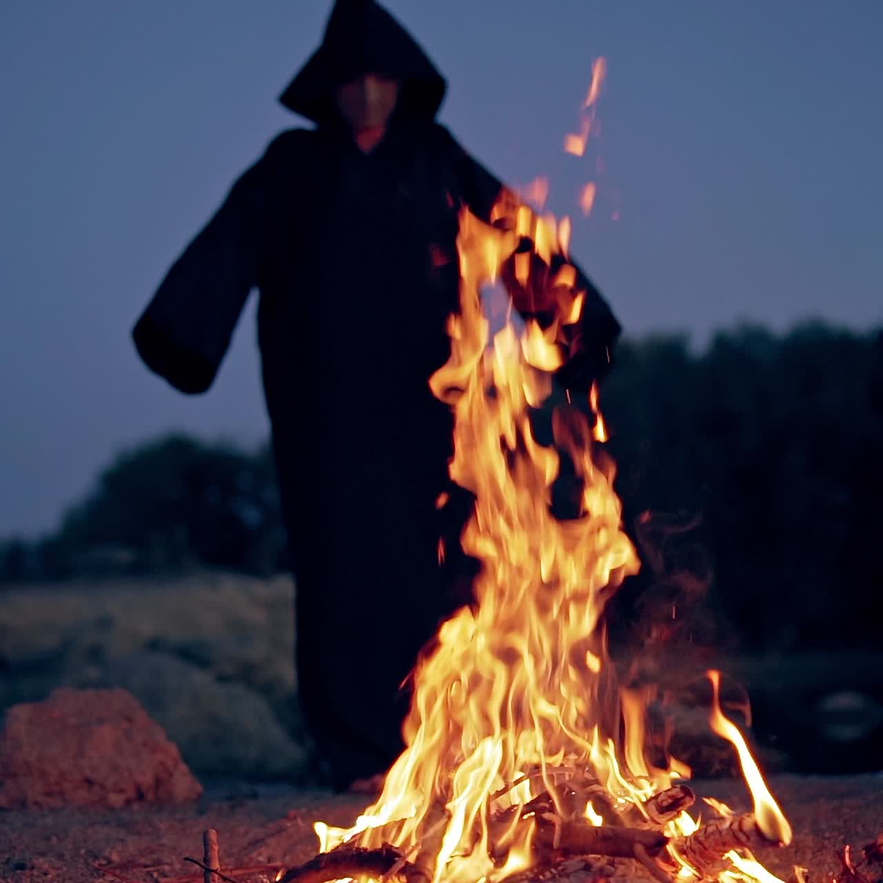 Fire and black figure at darkness. Spooky death in cloak with hood standing against the bright flame and doing some ritual outdoors. Halloween.