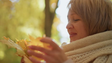 side view of woman wrapped in thick scarf smiling gently while admiring vibrant autumn leaf in hand as soft golden sunlight filters through trees creating dreamy seasonal glow