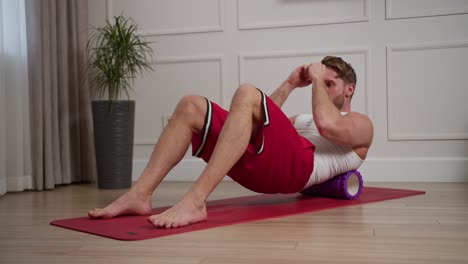 A-confident-blond-man-with-an-athletic-build-and-stubble-in-a-white-T-shirt-and-red-shorts-stretches-his-back-and-does-a-massage-with-a-massage-roller-during-his-morning-exercises-in-a-modern-bright-apartment-in-the-morning