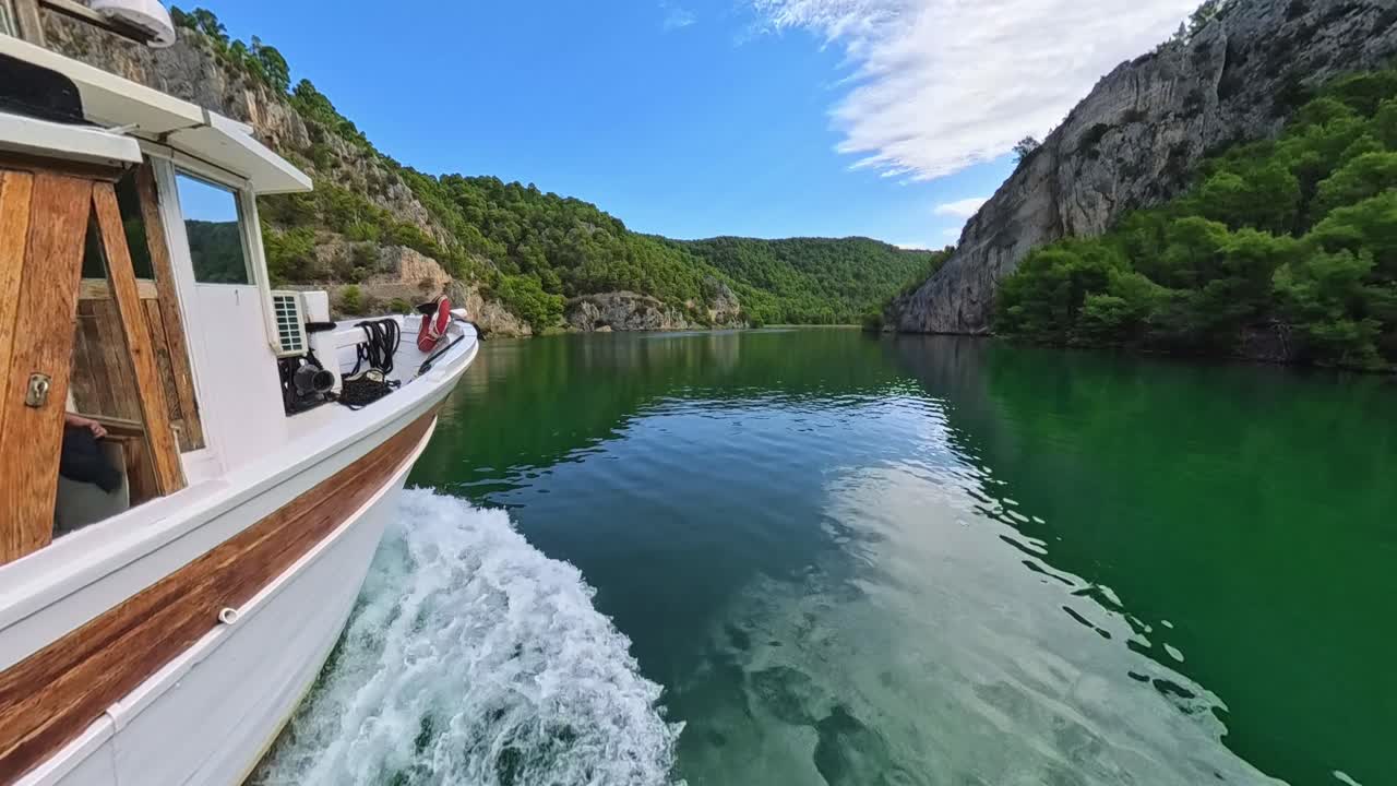 Footage from a moving tourist boat on the calm, green Krka River in Croatia. The view glides through a dramatic gorge with forested slopes on both sides, showcasing the park’s natural beauty