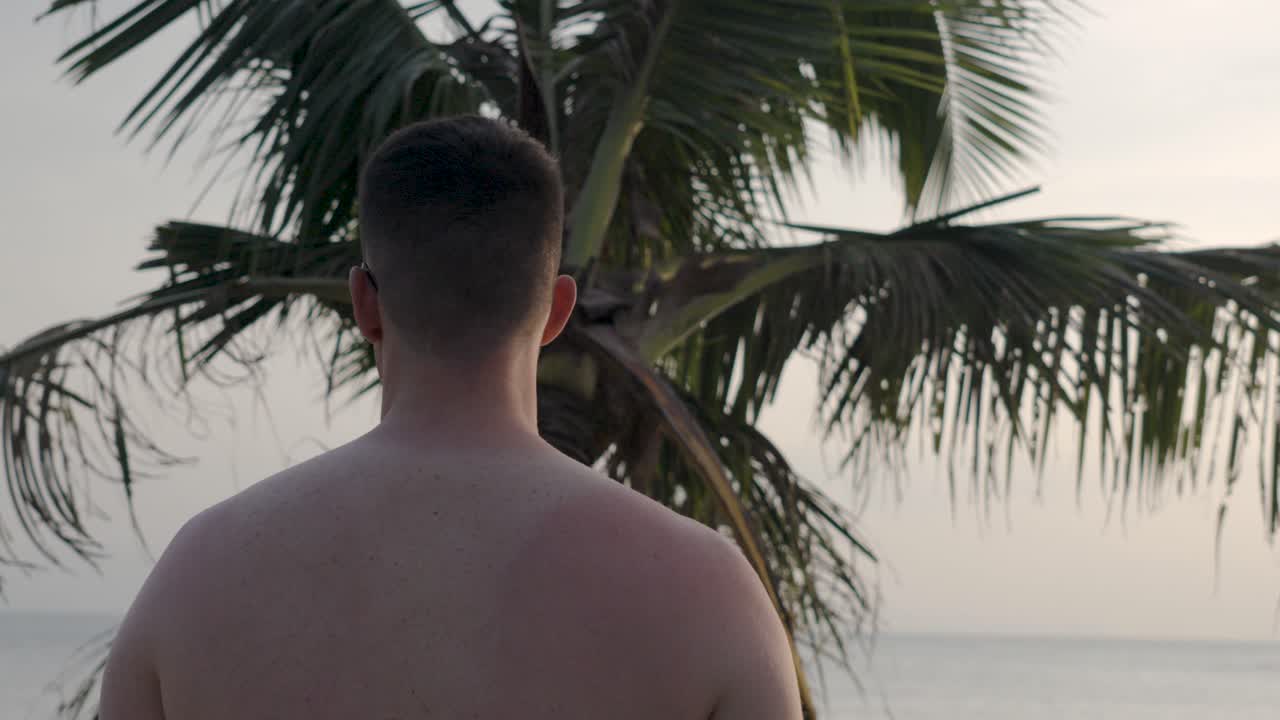 A man enjoys the peaceful tropical scenery on a beach in Koh Phangan, Thailand, surrounded by palm trees and ocean views.