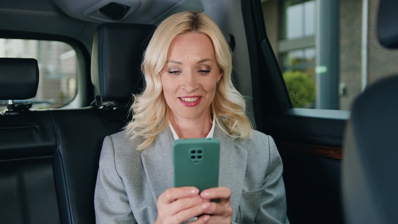 mujer sonriente montando en el asiento trasero del coche, escribiendo en el teléfono inteligente y mirando a través de la ventana