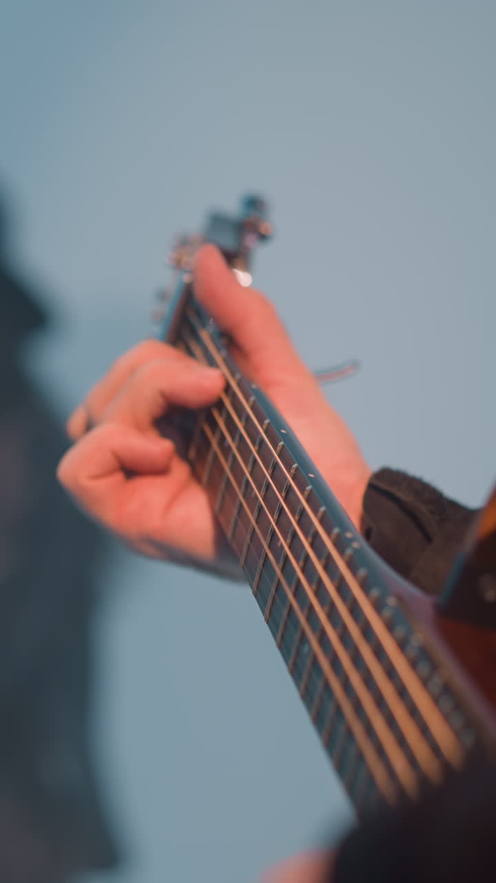 vista de cerca de un guitarrista sin rostro tocando una guitarra contra un fondo blanco con un cantante borroso y un tecladista en el fondo. la mano del guitárista está enfocada mientras tocan las cuerdas