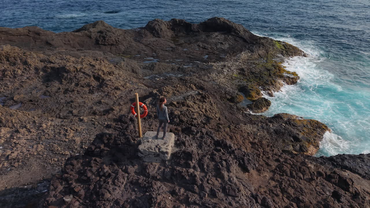 Scenic panoramic view capturing female tourist overlooking rugged volcanic coastline near historic Punta Sardina lighthouse with sweeping Atlantic Ocean horizon