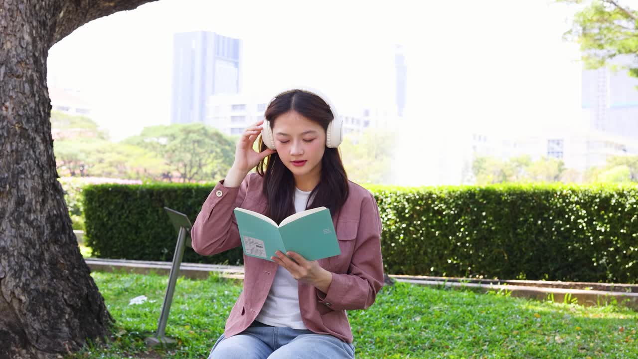 หญิงสาวนั่งอ่านหนังสืออยู่บนสนามหญ้าในมหาวิทยาลัยพร้อมกับสวมหูฟังสีขาว