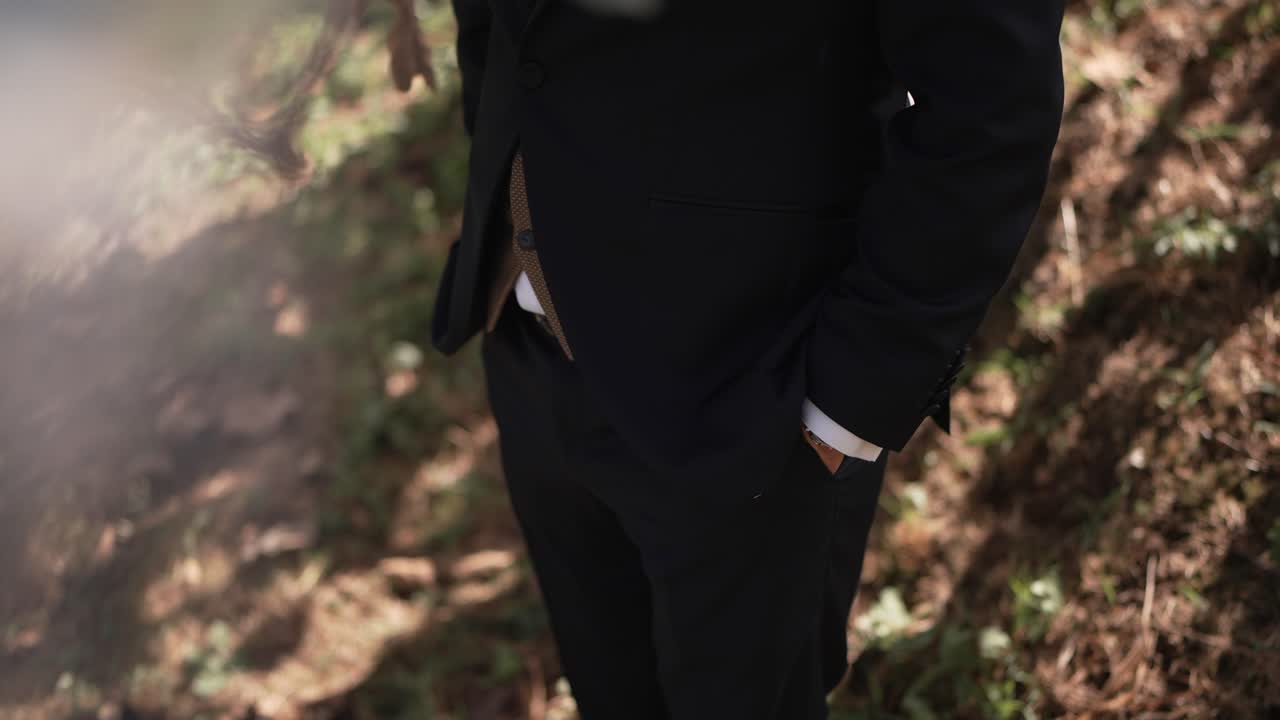 Close up of a groom in a black suit, hands in pockets, standing in a natural outdoor setting