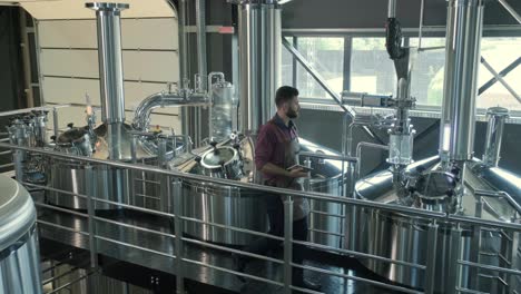 young male brewer wearing a leather apron supervise the process of beer fermentation at a modern brewery factory