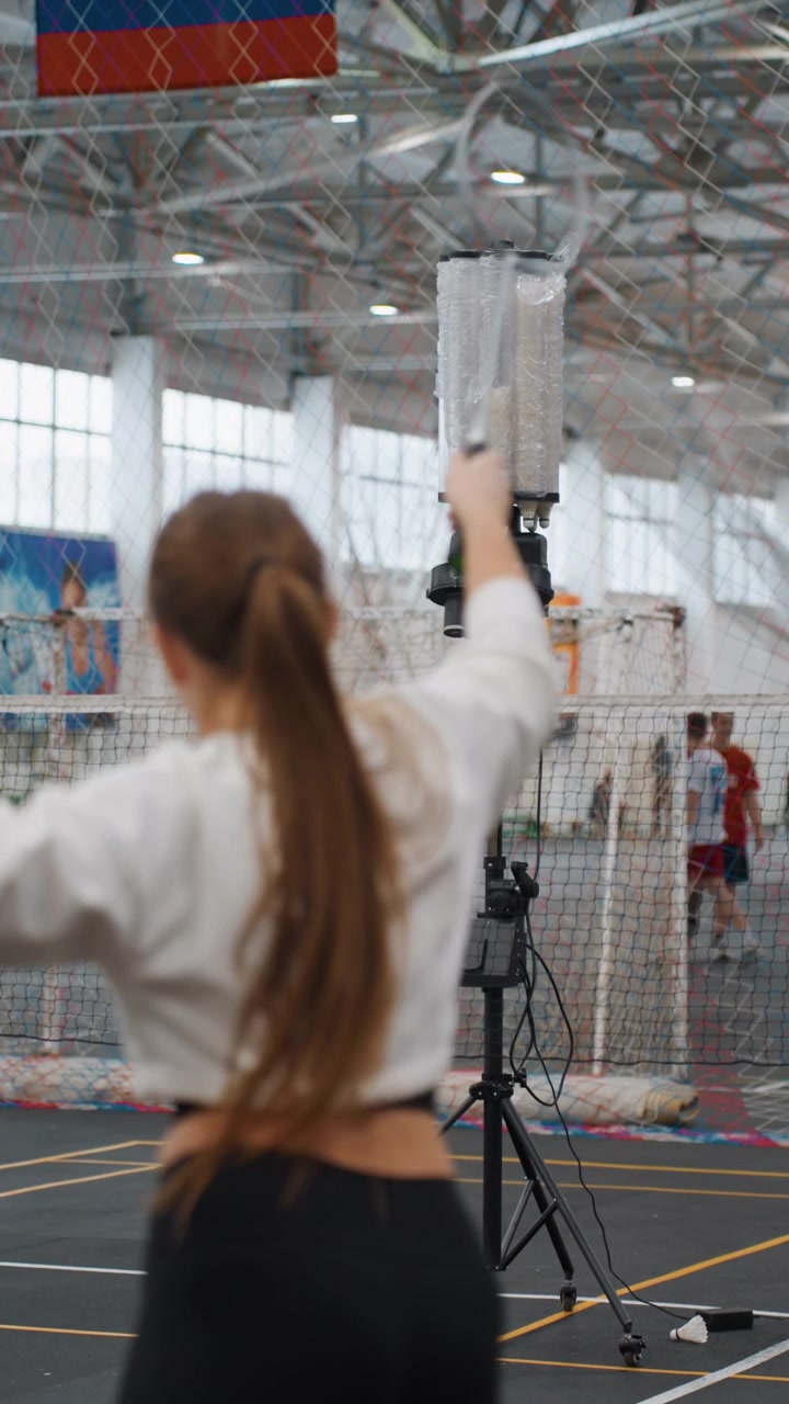 Trénink na badmintonovém kurtu v hale s podavačem míčků, hráčka zezadu, podávací stroj na stativu umístěný u sítě, trenér a spoluhráči rozmazaní v pozadí, osvětlení tělocvičny s vysokým stropem, neformální