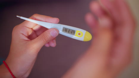 close up view of hands holding digital thermometer reading 41.1 degrees celsius while other hand raises partially covering display suggesting concern urgency and attention to high fever