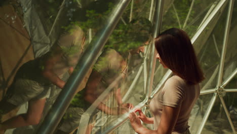 young lady looks out window examining area with dense forest. sun rays fall on female with reflection in panoramic window of modern apartment for tourists upper view