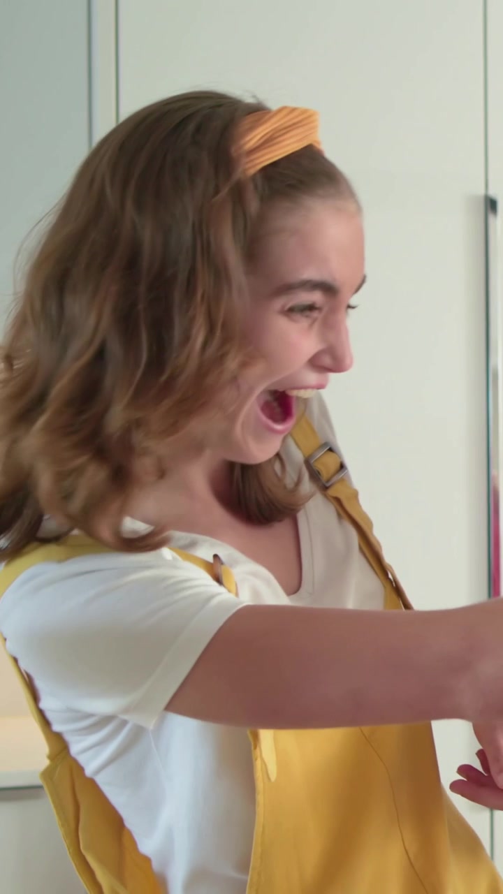 Vertical video: Laughing woman in yellow overalls scooping frosting in kitchen, decorating cake