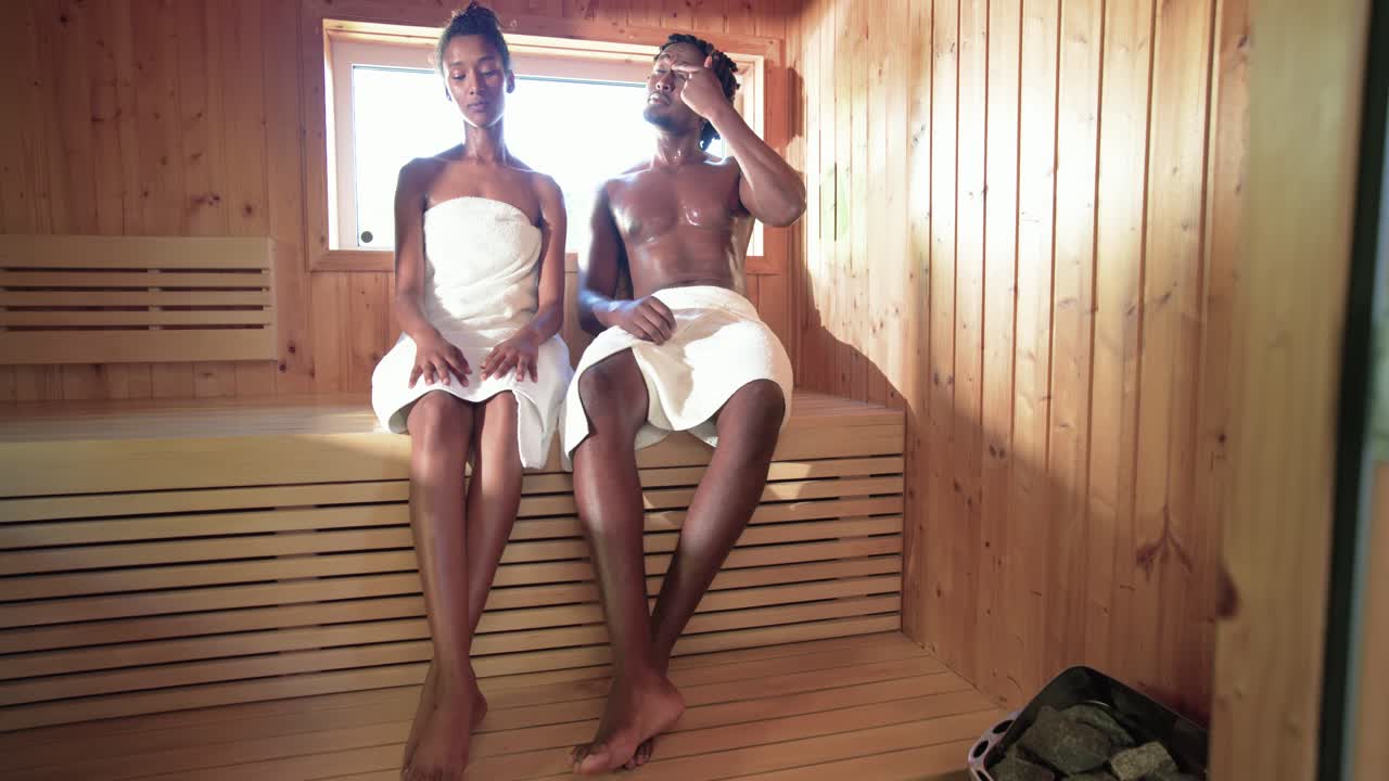 Casal afro-americano entrando na sauna e sentando no banco superior, ajustando toalhas, buscando calor