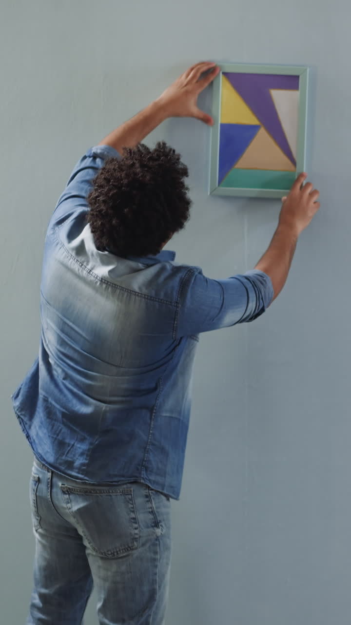 Black man chooses place to hang picture on wall with friend help in restored living room. Interior decorators work upon domestic decor. Home ornate