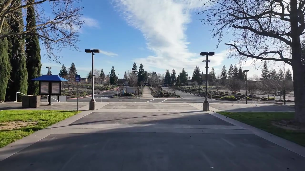 Sunlit wide angle shot of Cosumnes River College pathway and vacant parking lot in Sacramento. Shadows from trees and lampposts frame clear winter skies for serene, open campus footage.