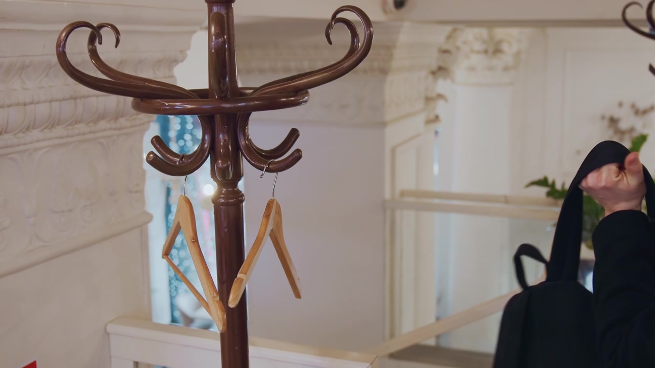 Un uomo in cappotto scuro appende il suo zaino di pelle nera a un gancio di legno intagliato di un supporto in metallo marrone, poi lo toglie e si allontana. La scena presenta il retro decorativo di una modanatura architettonica bianca