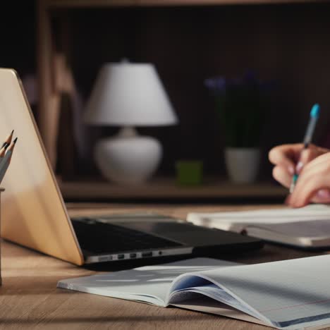 a woman's hand writes in a notebook sitting near a laptop at home