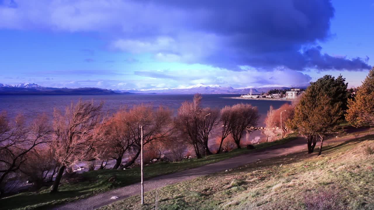 Uzaktaki Karlı Dağlar ve Dramatik Gökyüzü ile Manzaralı Göl Kenarı Manzarası.