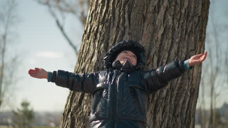 a young boy leaning his back against a large tree trunk, looking upward with a smile and his mouth open, both of his arms are raised up